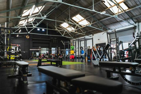 Placeholder photograph: a wide strength-training floor under a high roof, daylight coming through roof lights onto benches, racks and colour-coded plates receding down the room